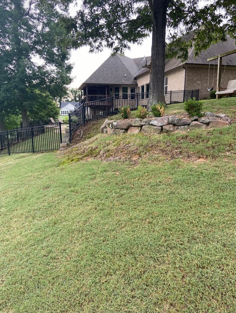 House with stone landscaping and black fence, surrounded by lush green grass and trees.