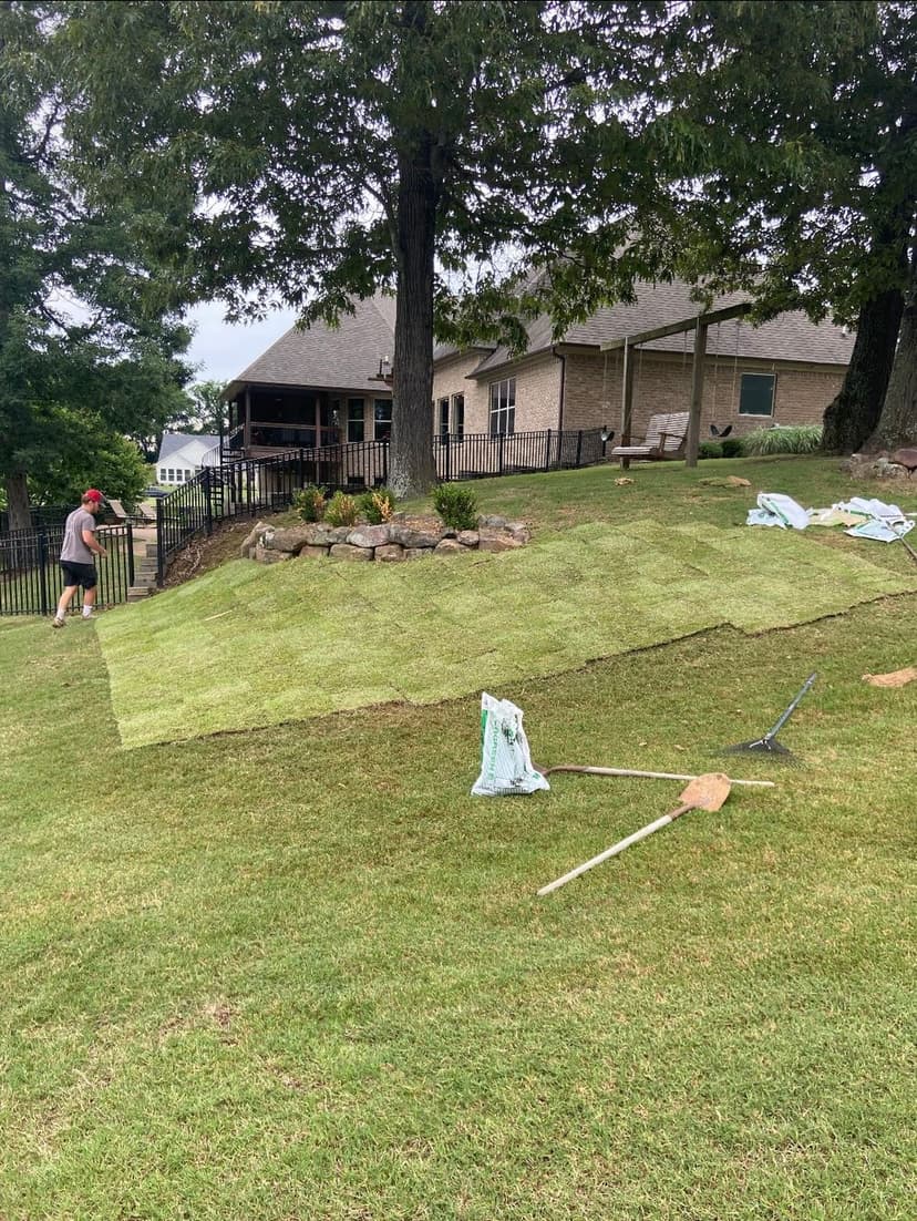 Lawn care professional installing fresh sod on a sloped yard near a house with trees.