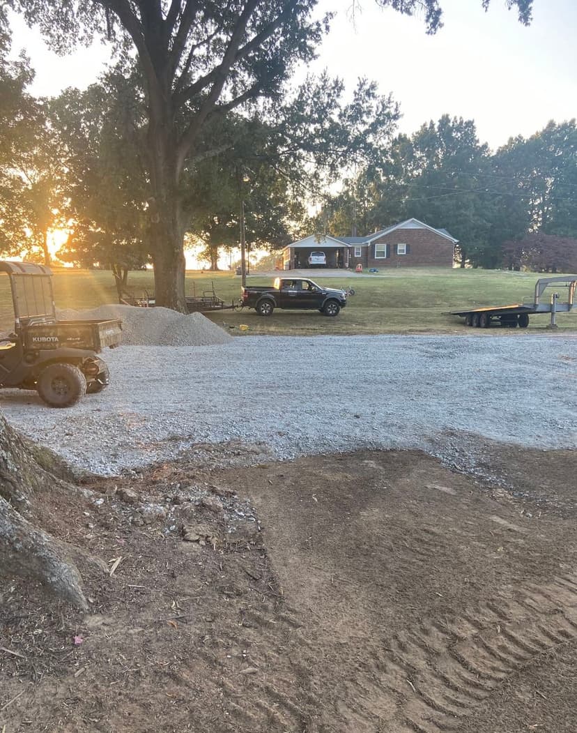 Gravel driveway under construction with a pickup truck and tractor near a house at sunset.
